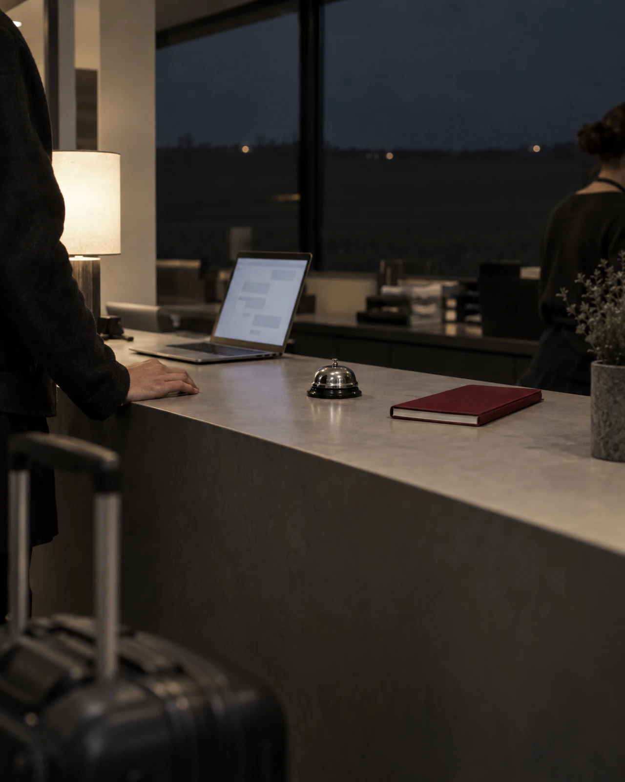 Hotelrezeption am Abend mit Laptop, Klingel und Notizbuch auf dem Tresen, davor ein Rollkoffer