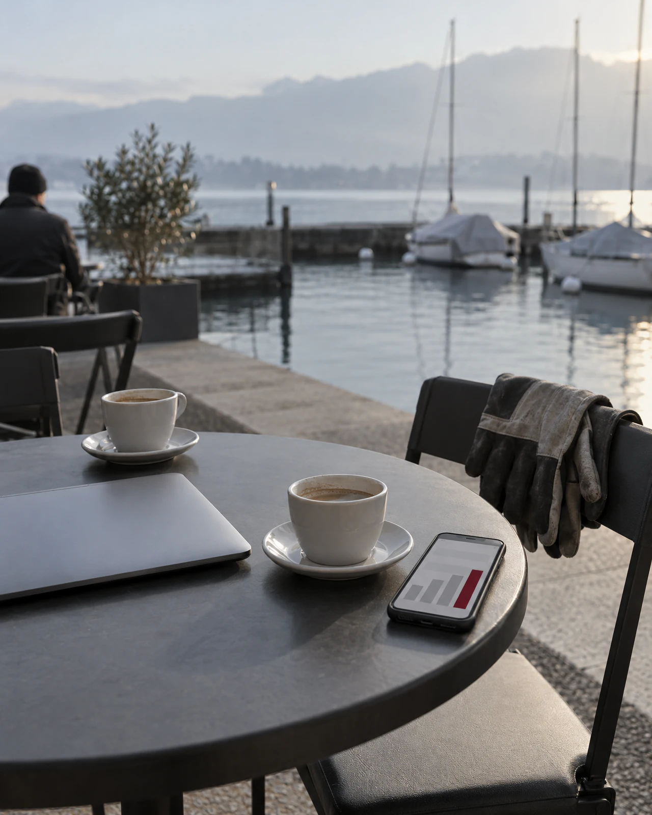 Zwei Kaffeetassen, geschlossener Laptop und Smartphone mit Balkendiagramm auf einem Tisch an einem Hafenbecken